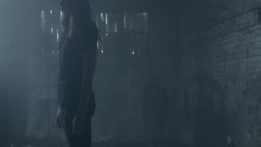 A young woman is standing still in a room with boarded up windows and glass on the floor