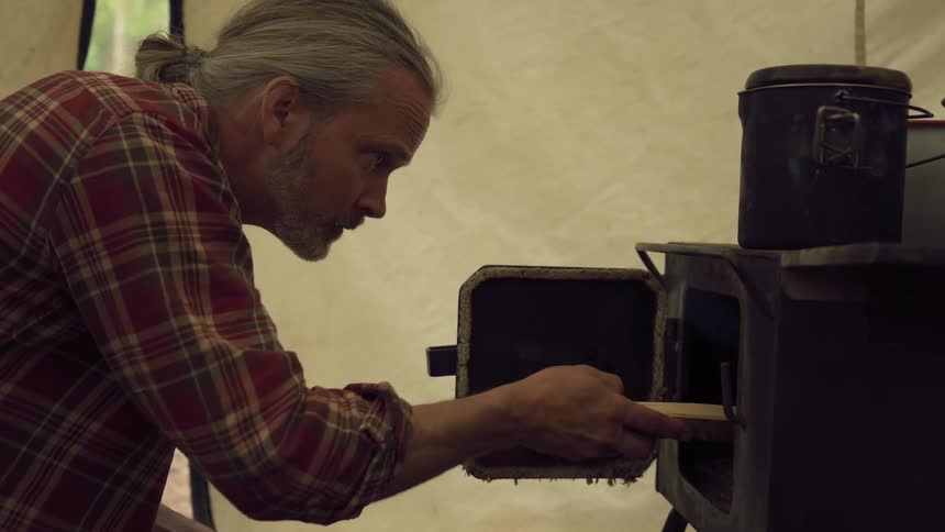 A man with grey hair tied back feeds a piece of wood into a small wood burning stove inside a canvas tent and blows on it