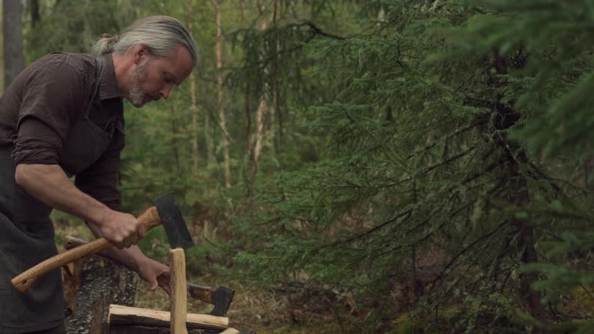 A whitehaired man chops wood with a small axe in a forest clearing