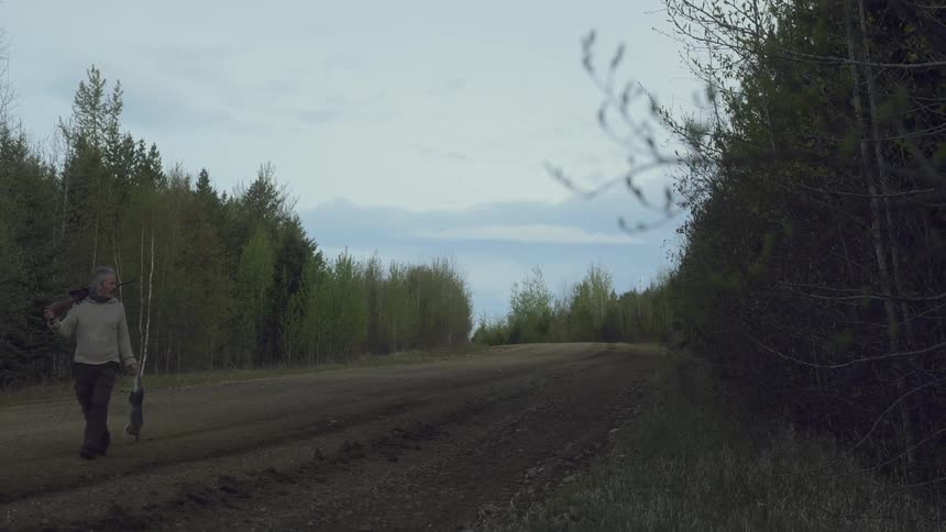 A hunter walks on the large dirt trail carrying his rifle and a dead rabbit