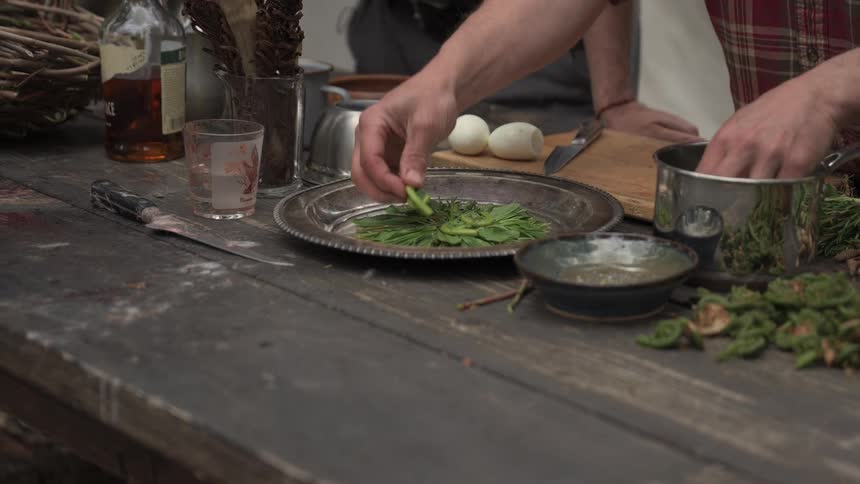 A person prepares food on a rustic wooden table surrounded by fiddlehead ferns boiled eggs and kitchen utensils