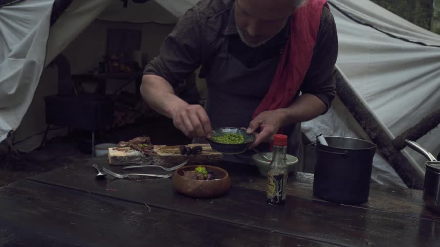 A grayhaired person adds green peas to a bowl of food on a wooden surface near a tent in a forest