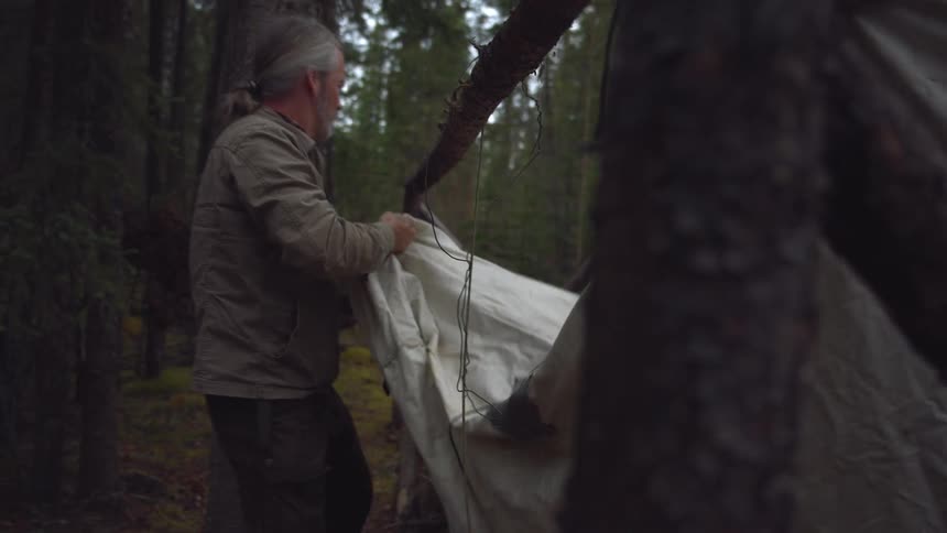 A man sets up a large tent pulling a tarp taut over a frame of logs in a forest clearing