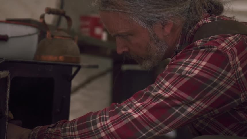 Older man lights a wood burning stove while in a canvas tent