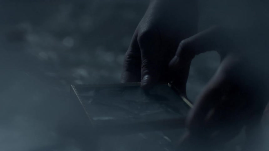 A hand reaches down to pick up a broken picture frame from a pile of glass