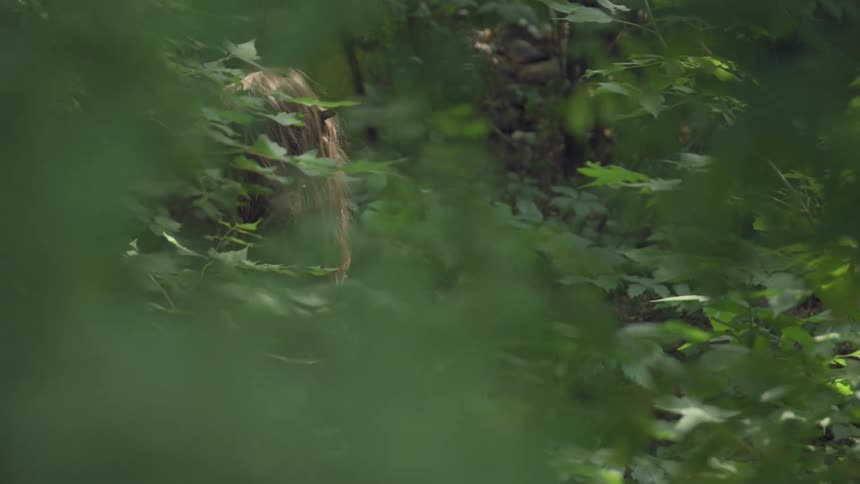 A girl with long brown hair is walking through a dense forest mostly hidden behind thick green leaves and branches with only part of her head and arm visible through the foliage