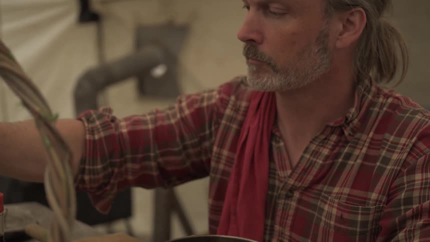 A person wearing a plaid shirt pours a dark liquid from a bottle into a pot on a wooden table in a rustic kitchen