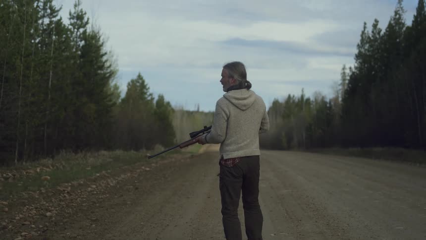 A hunter with long gray hair wearing a gray hoodie and dark pants walks along a dirt road carrying a rifle