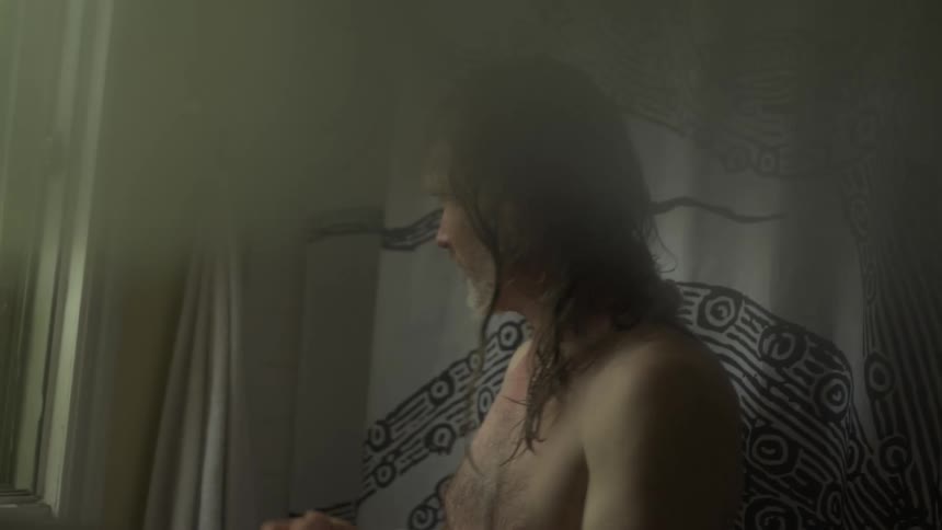 A man with long hair and a towel stands barechested in a bathroom his body and hair wet