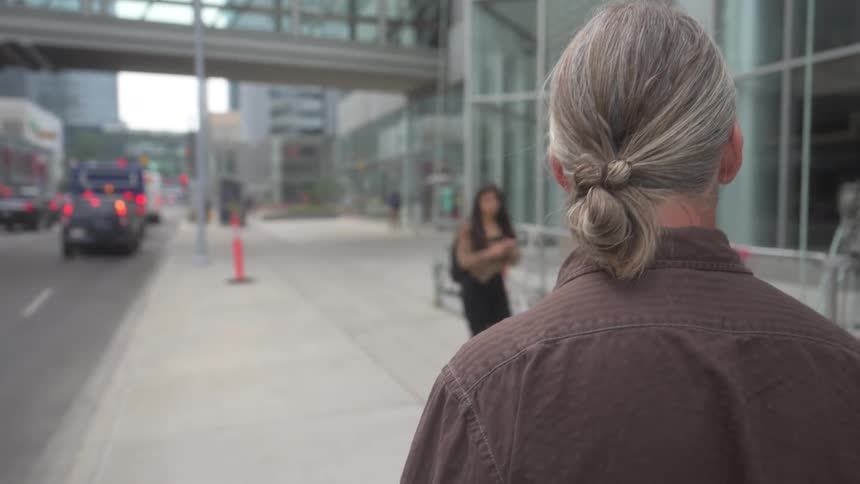 A person with gray hair pulled back walks along a city sidewalk next to a street