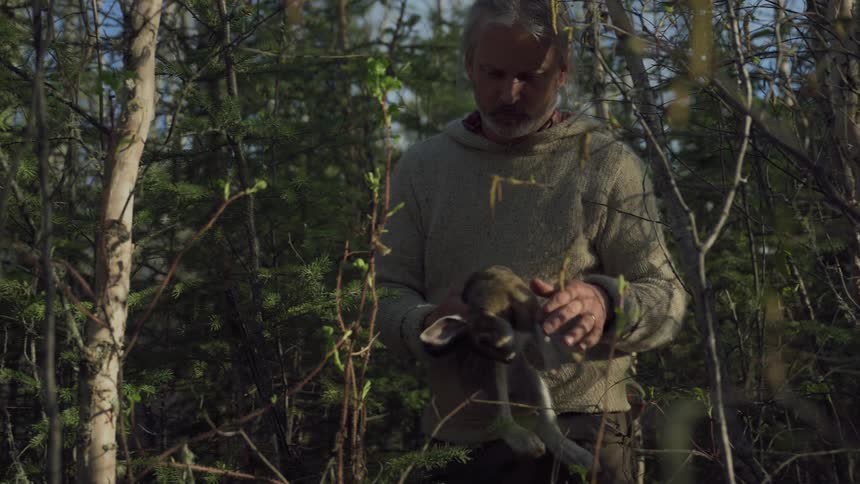 A man in a light colored sweater holds a dead rabbit in a wooded area with branches and foliage visible around him
