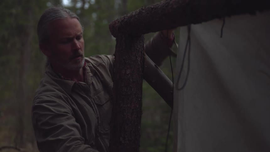 Older man uses cord to attach canvas onto a frame formed of logs while camping in the woods