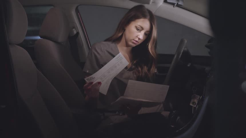 A woman with long brown hair sits in the drivers seat of a car at night illuminated by the interior light and reads a document
