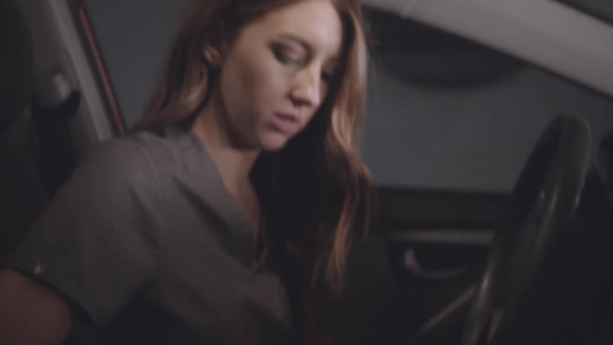 A woman with long brown hair sits in the drivers seat of a car at night illuminated by the interior light and reads through papers