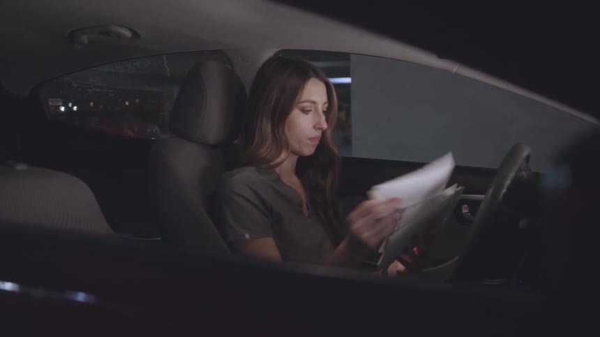 A woman with long brown hair sits in the drivers seat of a car at night illuminated by the interior light and reads through papers