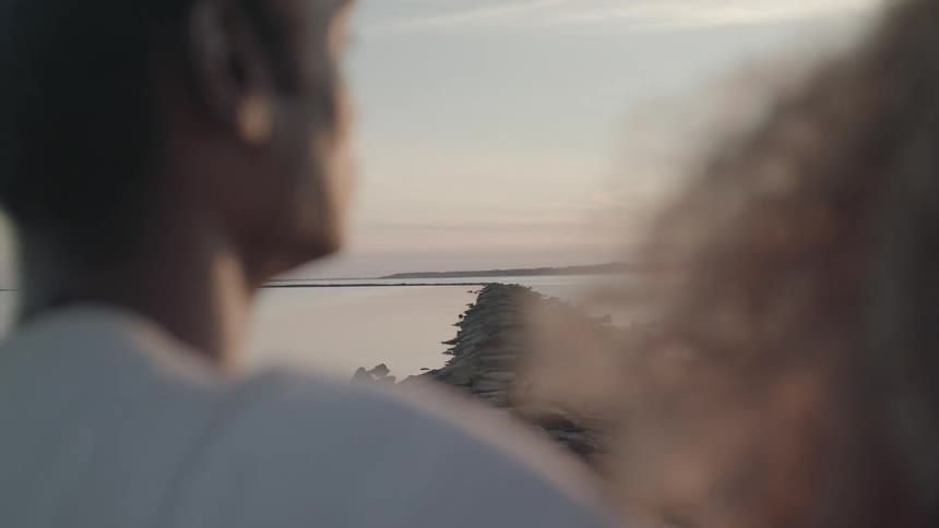 Two people stand near a long narrow rock jetty stretching into the water at sunset