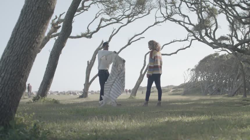 A young couple lies on a blanket spread over grass in a small grove surrounded by trees on a calm morning