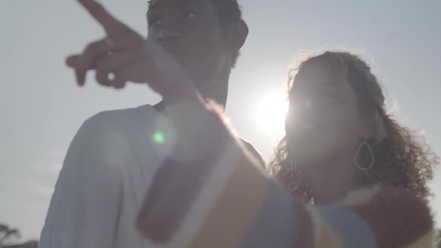 A teenage girl in a colorful striped sweater gestures toward something while a young man in a white shirt stands nearby with bright sunlight behind them
