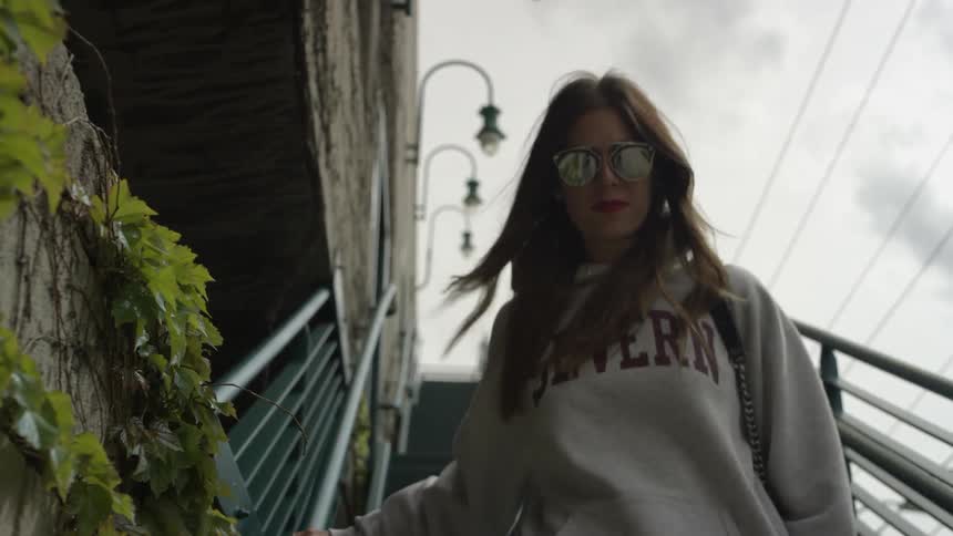 Woman wearing a gray hoodie and sunglasses walking down stairs in a city on a cloudy day