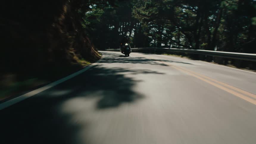 A motorcycle is seen driving away on a paved road curving through a tree lined landscape