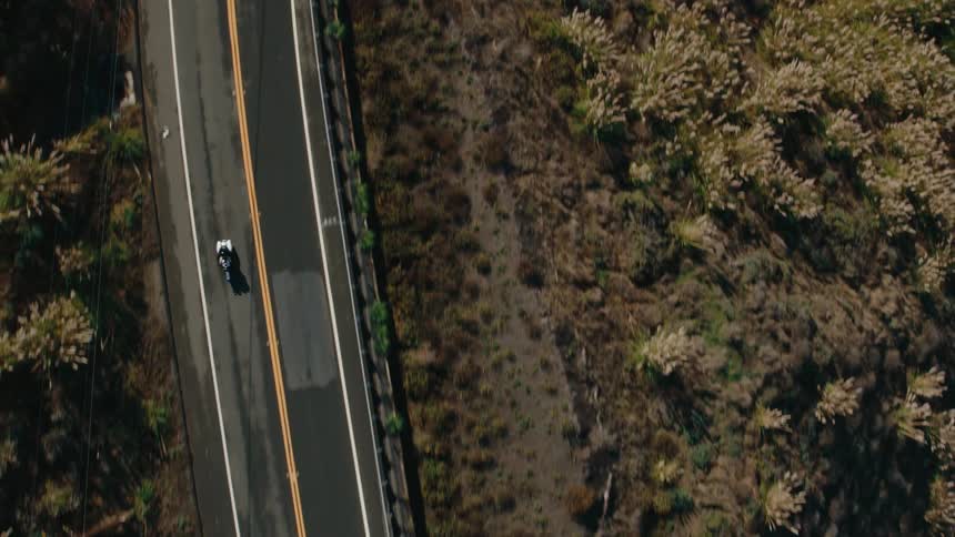 An aerial view of a motorcycle navigating a sharp curve on a coastal highway with the road winding along a rugged hillside overlooking the ocean