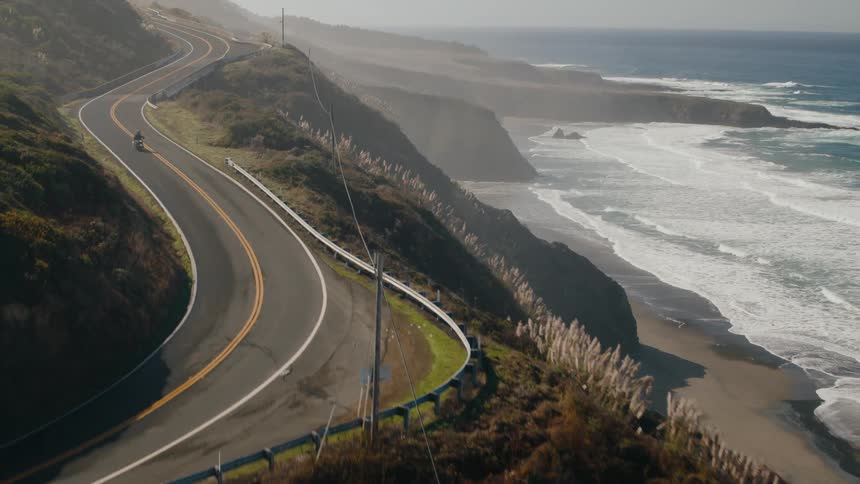 The motorcycle drives on the road by a cliff near the beach