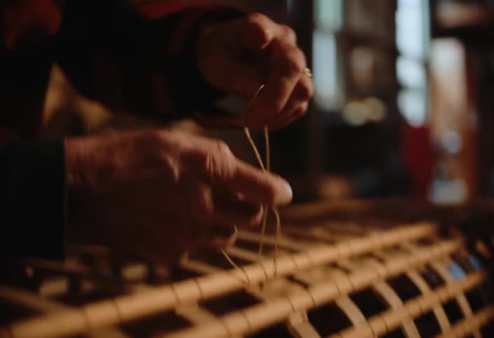 A bearded man wearing a hat and glasses works on a handcrafted structure in a dimly lit wooden workshop