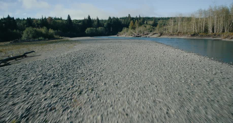 A wide shallow river flows through a gravelly riverbed flanked by trees and vegetation under a clear sky