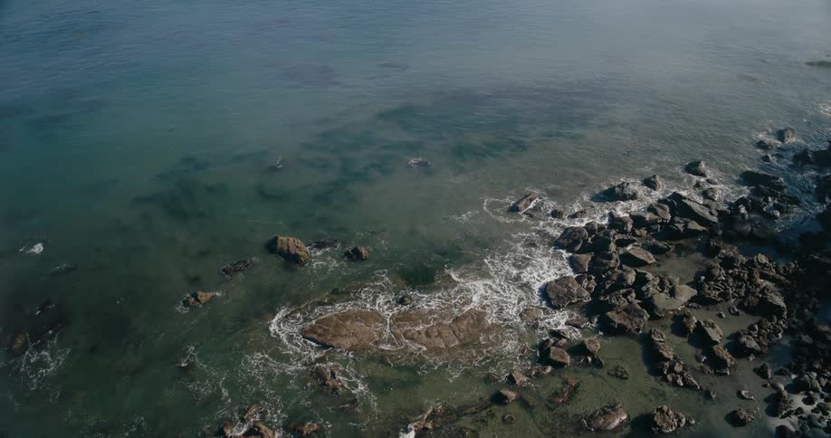 An aerial camera glides toward a rugged coastline capturing rocky outcrops and clear shallow waters where the sea gently meets the shore