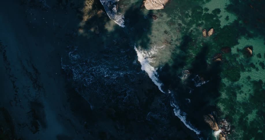 A rocky shoreline where the dark jagged rocks meet the clear turquoise waters of the ocean with white waves crashing against the rocks