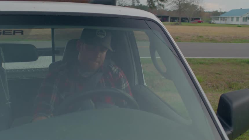 A man wearing a plaid shirt and hat drives a truck along a country road parks at his home and sighs with exhaustion