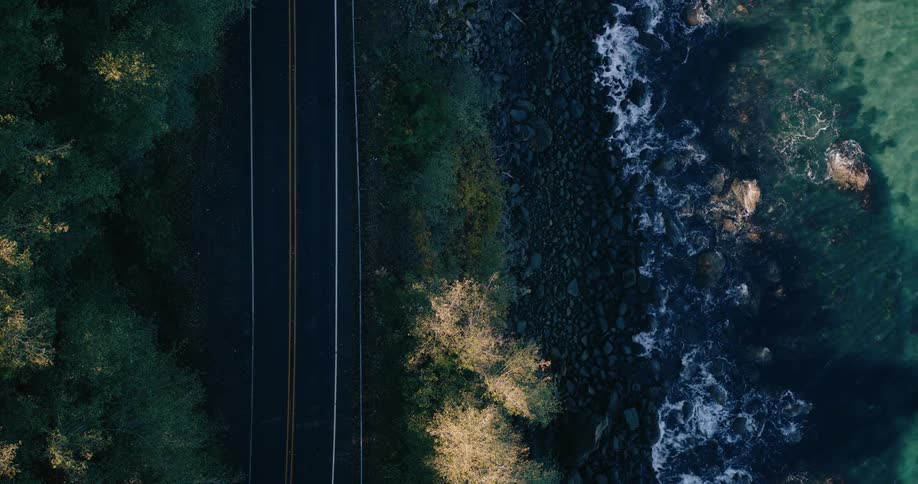An aerial view of a straight road running parallel to a rocky shoreline where the dark rugged rocks meet the clear turquoise waters of the ocean