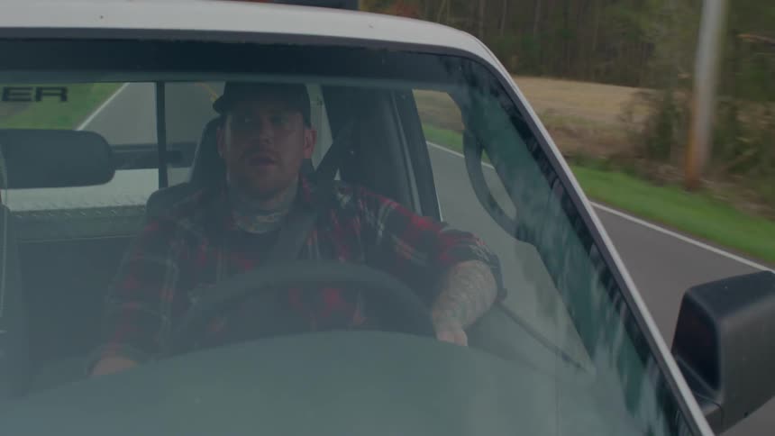 A man wearing a plaid shirt and a hat drives a car or truck along a countryside road