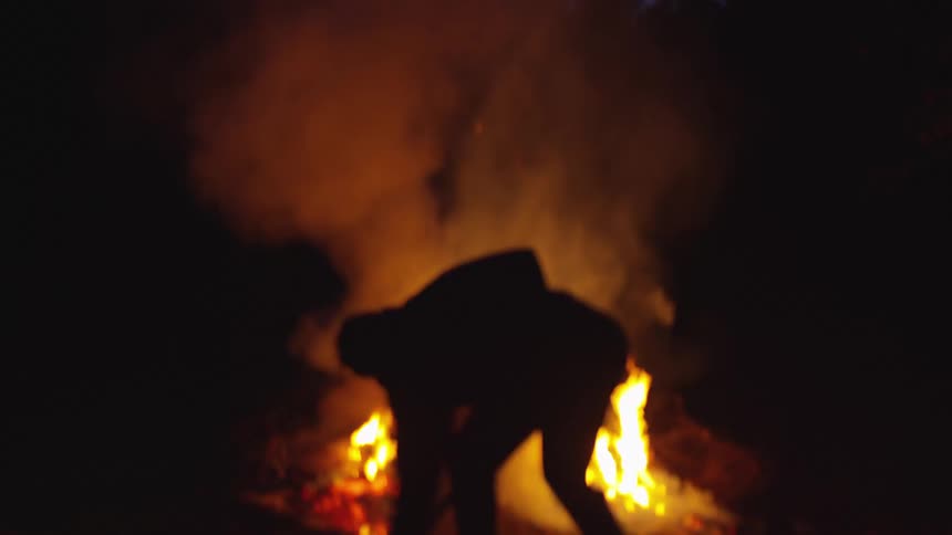 A shadowy silhouette desperately shovels dirt into a blazing bonfire the flickering flames casting stark contrasts against the surrounding darkness