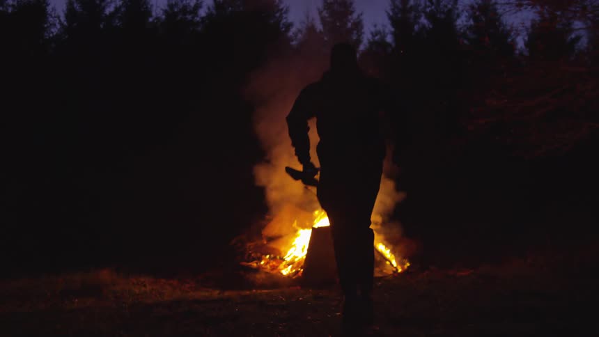 Silhouette of a man running towards a bonfire in the middle of a forest at night
