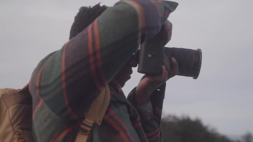 A man with short dark hair wearing a patterned jacket and a backpack looks through the viewfinder of a camera