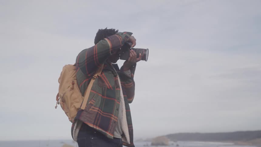 A man with dark hair wearing a plaid shirt and a backpack holds a camera to his eye taking a photograph of the scenery