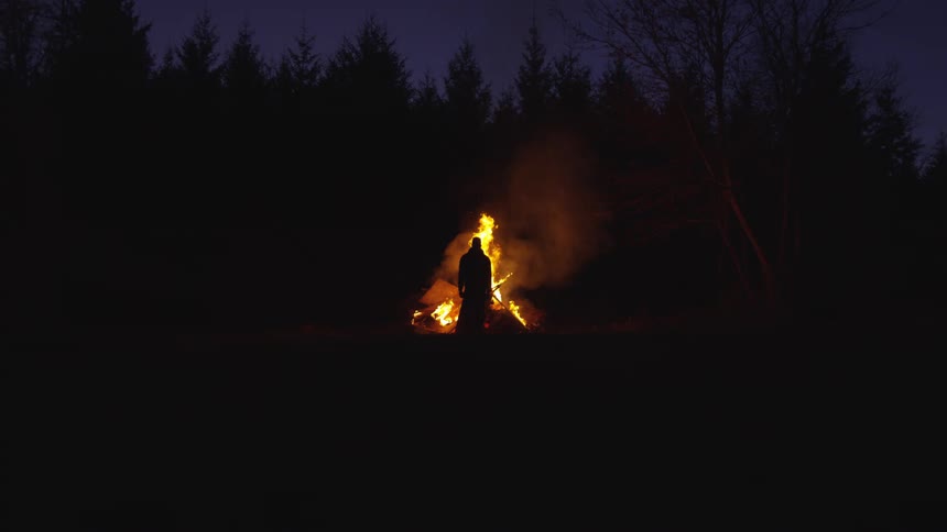 A silhouetted person stands before a large bright bonfire burning in an outdoor area near trees at night