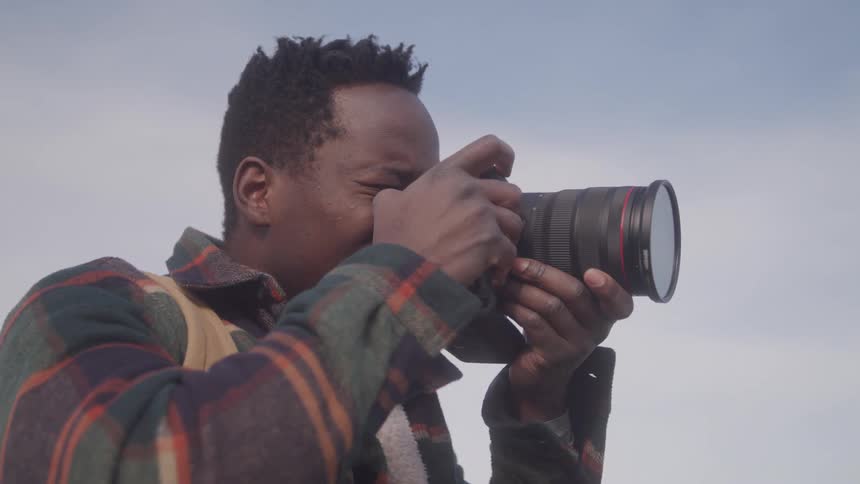 A man with short dark textured hair wearing a patterned shirt focuses intently as he holds a professional camera to his eye suggesting the act of capturing a photograph
