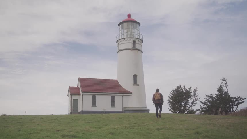 A lone traveler with a backpack walks across a grassy field toward a tall coastal lighthouse under a soft cloudy sky