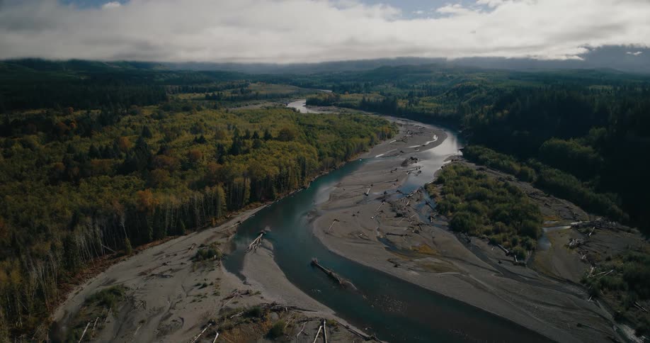 An aerial view of a wide river winding through a lush forested valley with gravel bars and driftwood visible along its banks under a partly cloudy sky
