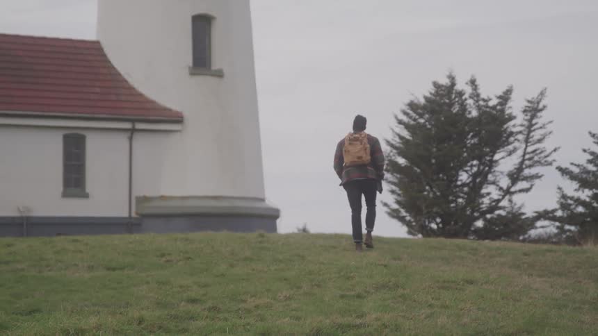 A person wearing a backpack walks up a grassy hill toward the base of a white lighthouse building on an overcast day
