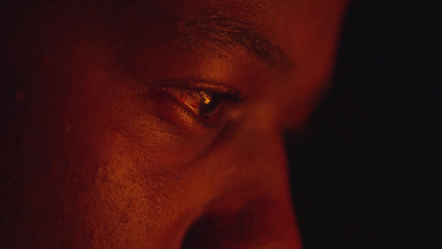 A mans eye reflecting a warm reddish light source with visible skin texture and eyebrow details against a dark background