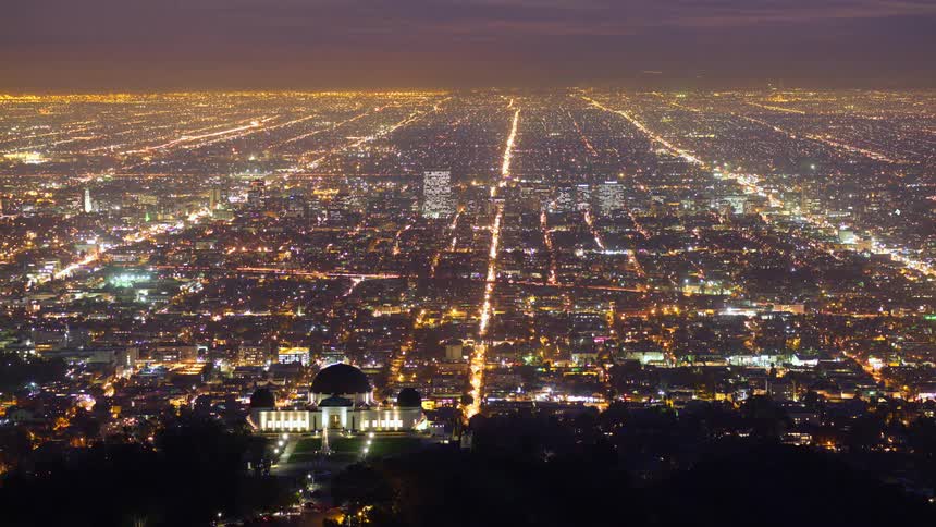 An expansive aerial view of the twinkling lights of los angeles at dusk with the griffith observatory prominently featured on a hilltop overlooking the gridlike pattern of the city streets stretching into the distance