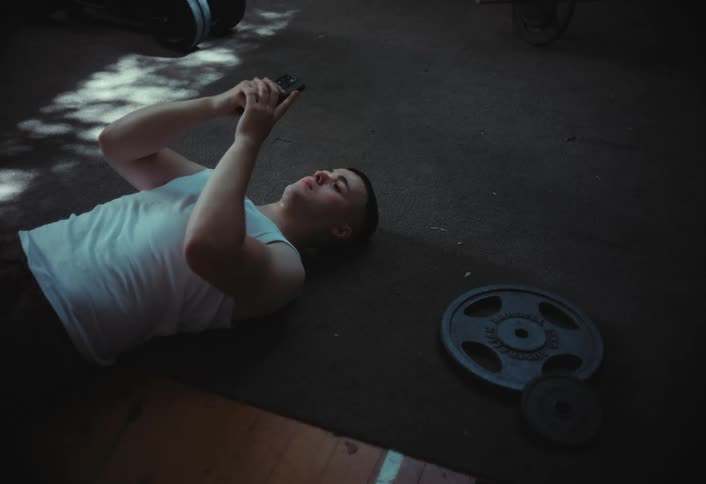 A person lies on the floor of a gym holding a smartphone above their face surrounded by weight plates and fitness equipment