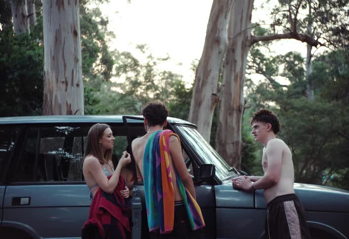 Three young people two shirtless males and a female in a swimsuit stand around a teal suv in a wooded area after swimming as two of them are draped in towels