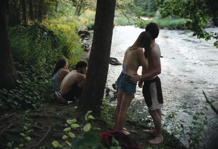 Teenage couples are hanging out next to a river as one couple kisses