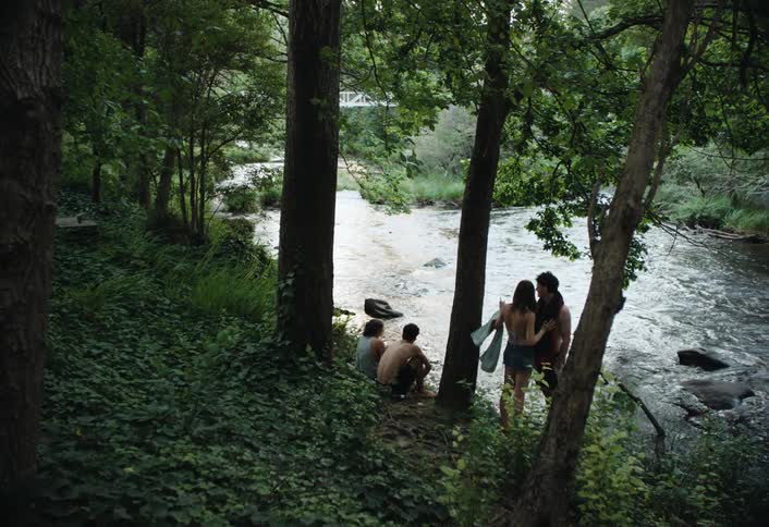 A group of four people near a flowing river surrounded by trees and lush greenery
