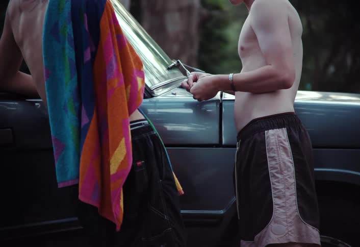 Two young males likely teenagers are having a conversation near a blue truck both wearing board shorts and no shirt with one removing a colorful beach towel from his shoulder to reveal tattoos along the length of his shoulder blades