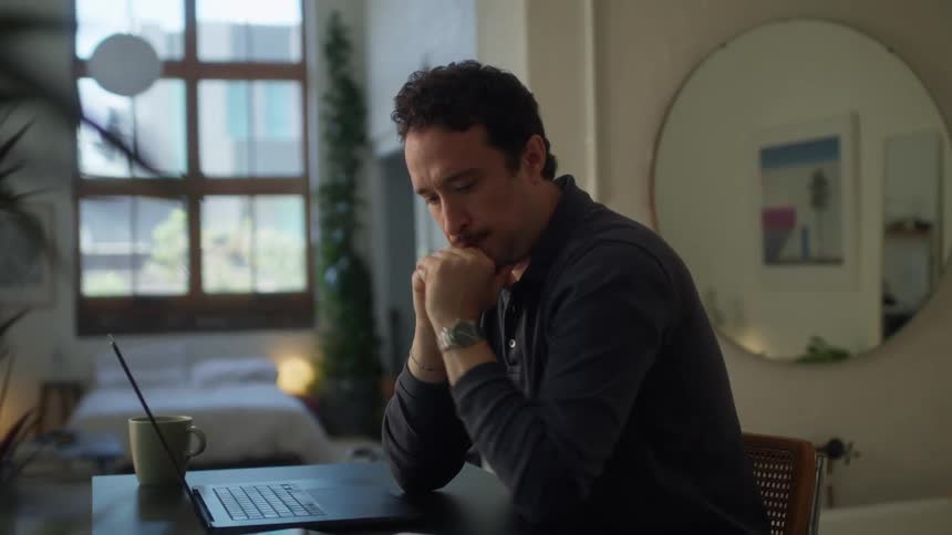 A man with dark curly hair sits at a desk in a well lit room focused on typing on a laptop